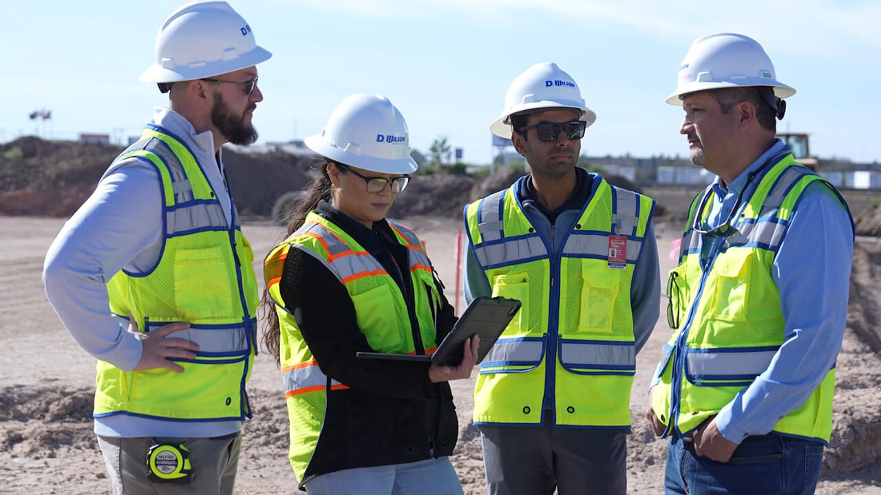 Four D Wilson employees standing together, discussing a project with a construction site behind them.